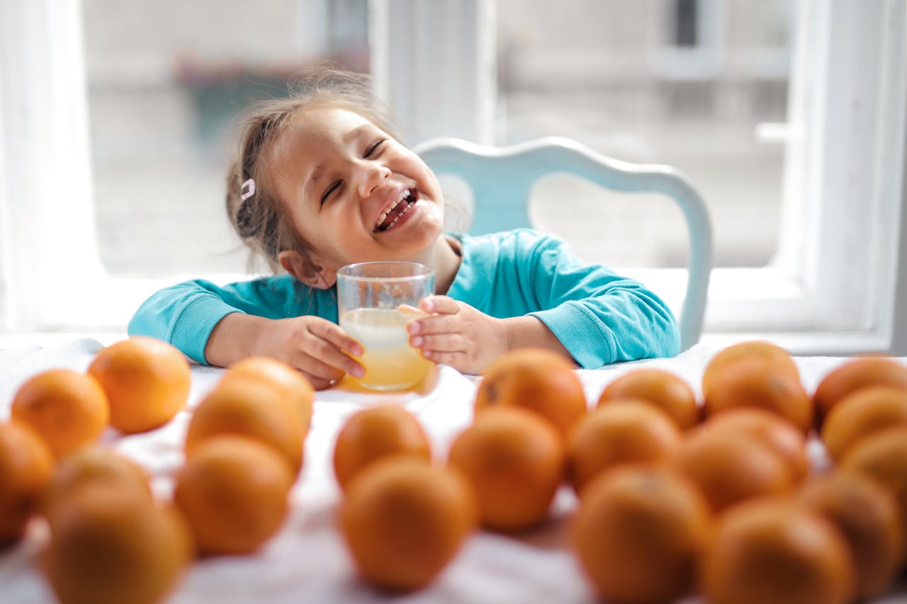 A cheerful child smiling while holding a glass of fresh orange juice surrounded by oranges indoors.